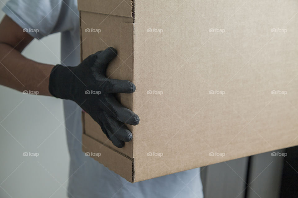 Detail of man with black gloves holding a cardboard box for delivering products on gray background. Delivery concept. Delivery service concept. Copy space.
