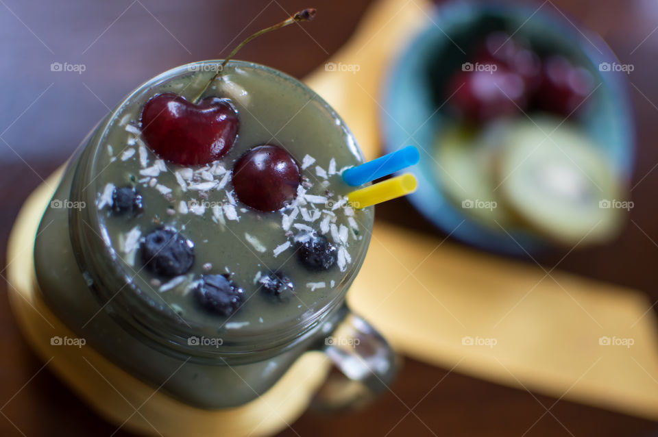 Fresh fruit garnish on green smoothie high angle view with black cherry topping, coconut and blueberry smile high angle view of smoothie in glass with drinking straws and kiwi and cherries in background on dark wood table part of a series