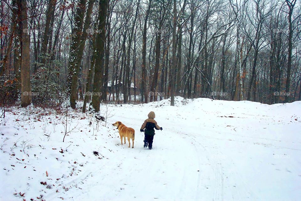 A young boy is seen running down a path on a snowy day with a Labrador retriever.