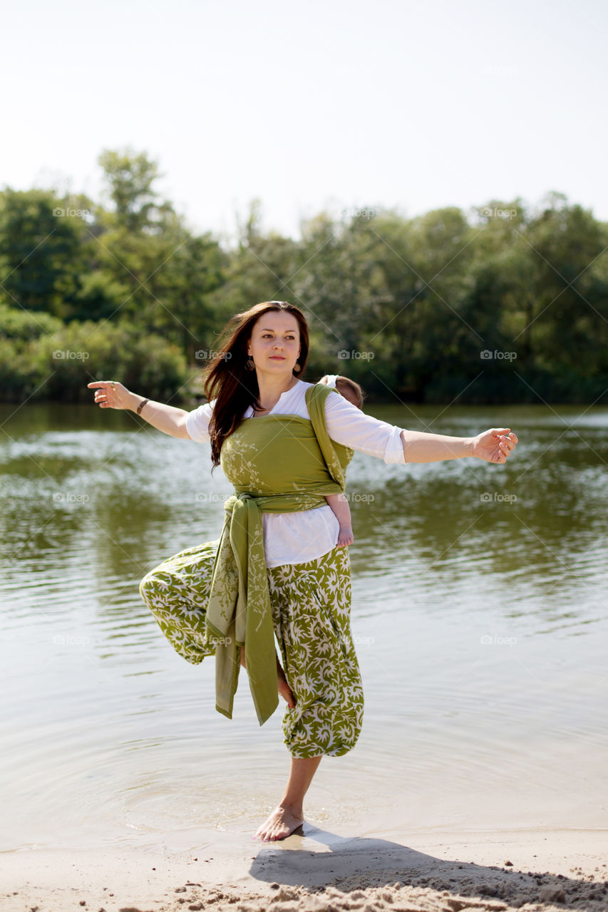 Mother doing yours at river beach with baby in sling 