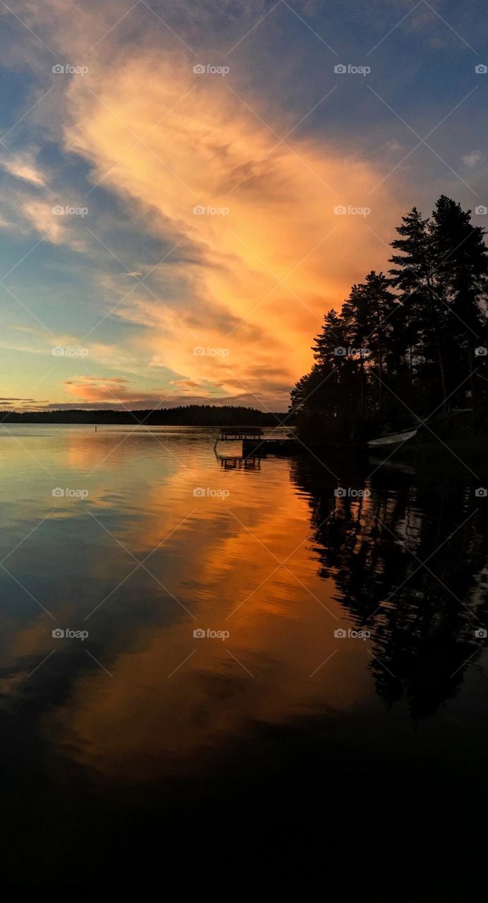 Amazing orange sunset over the horizon with dark trees and pier silhouette and the reflection in the smooth water surface 