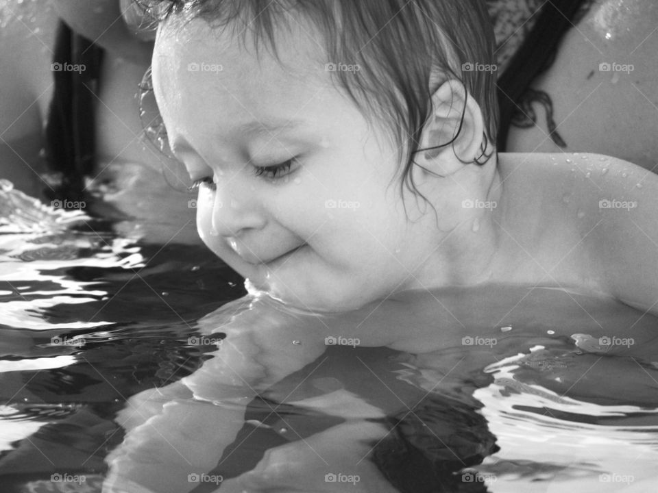 A small boy reaches for a submerged ring in a swimming pool from the safety of his mother’s arms. 