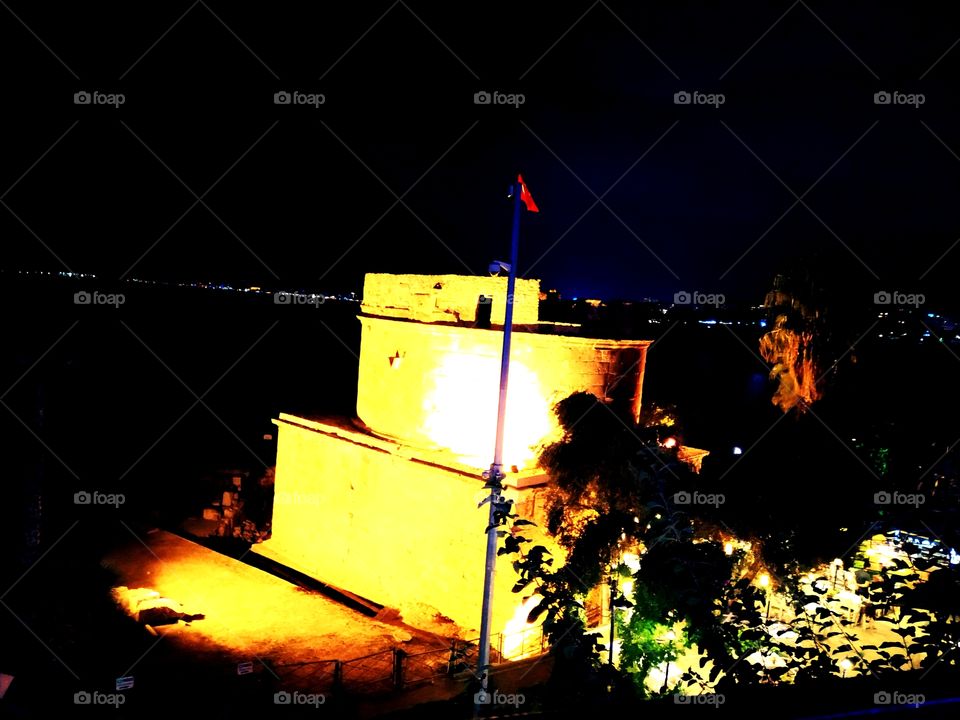 Artificially iluminated old fortress with white lights, nature in front and iluminated city far behind.