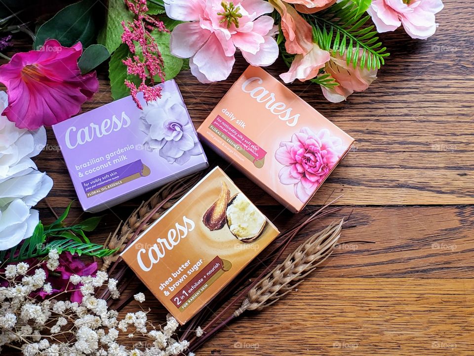 Left side oriented:  The new look Caress bar single packs:  Daily silk,  shea butter and brown sugar,  and Brazilian gardenia & coconut milk on a wood surface surrounded by matching florals.