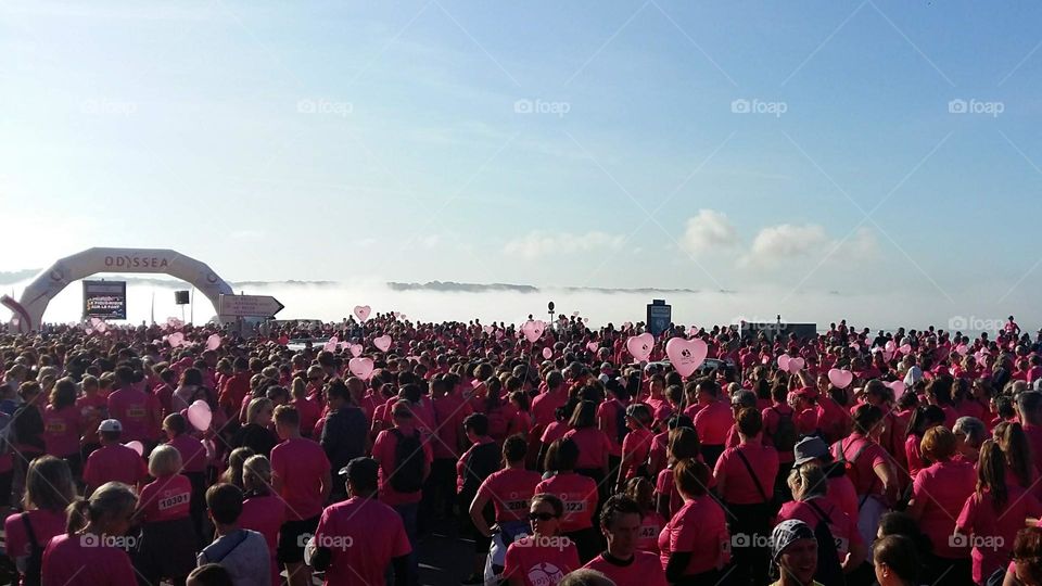 A crowd of athletes before the start of a race for breast cancer research. Everyone is wearing a pink t-shirt, some are racing with heart-shaped balloons.