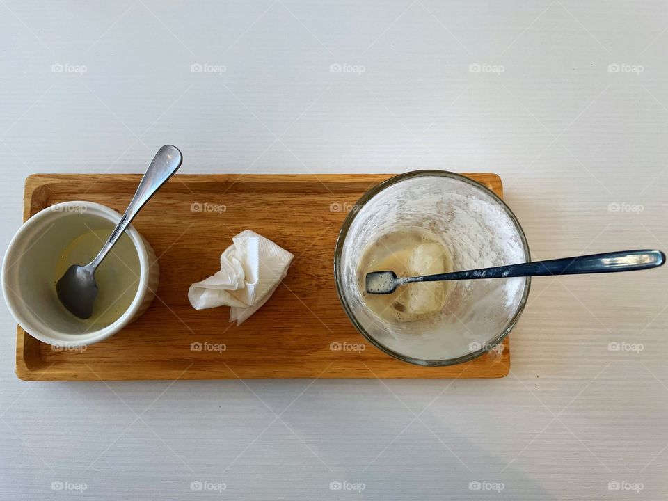 An empty latte with a black spoon sticking out, a crumpled tissues, and an empty ice cream cup with a silver spoon sticking out. This is the remnants of an latte float. The items are on a small wooden tray. 
