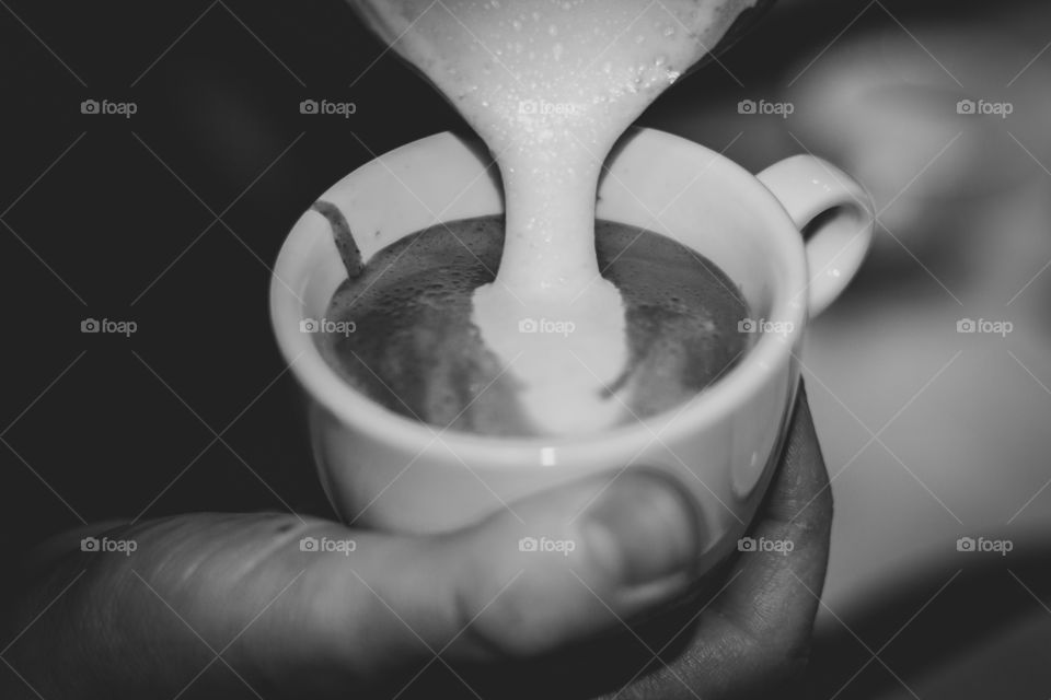 a black and white portrait of a coffee mug full of coffee being held by a barista pouring some milk into it.