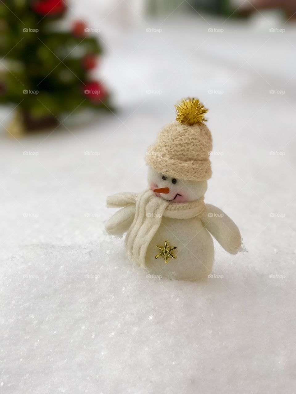 little snowman standing in the snow, behind a blurred background with a festive Christmas tree