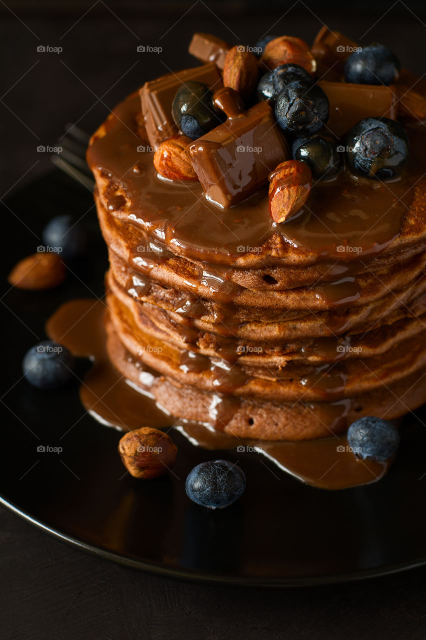 Stack of pancakes with chocolate sauce, blueberries and nuts on dark wooden desk