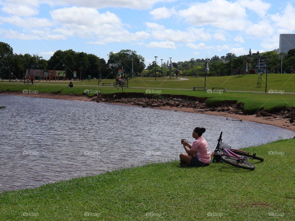 A young woman sitting on the edge of a serene lake in a park, absorbing the relaxing environment and enjoying a moment of tranquility. In the background, cyclists pass on a sunny day with white clouds in the sky