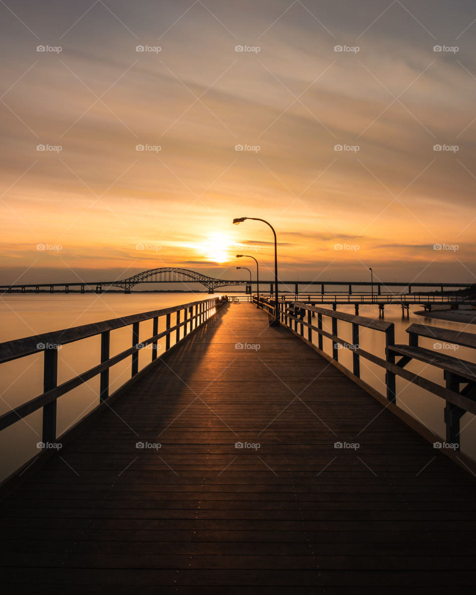 Wooden fishing pier leading out into the sunsets orange sunset colors reflecting on the water and the boardwalk. 