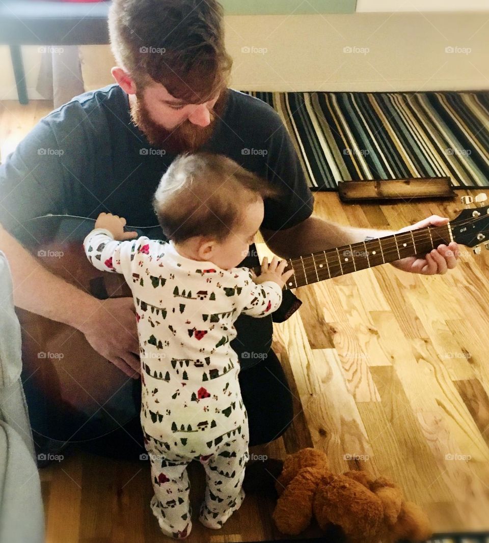 A young man gently plays the acoustic guitar for a small child in pajamas 