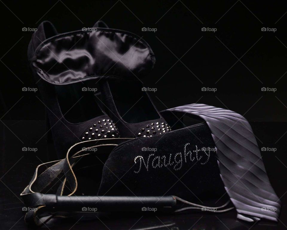 black high heels with silver accents draped with a black blindfold, two tone gray tie draped over a black handbag with the word naughty spelled in jewels and a black leather whip coming out