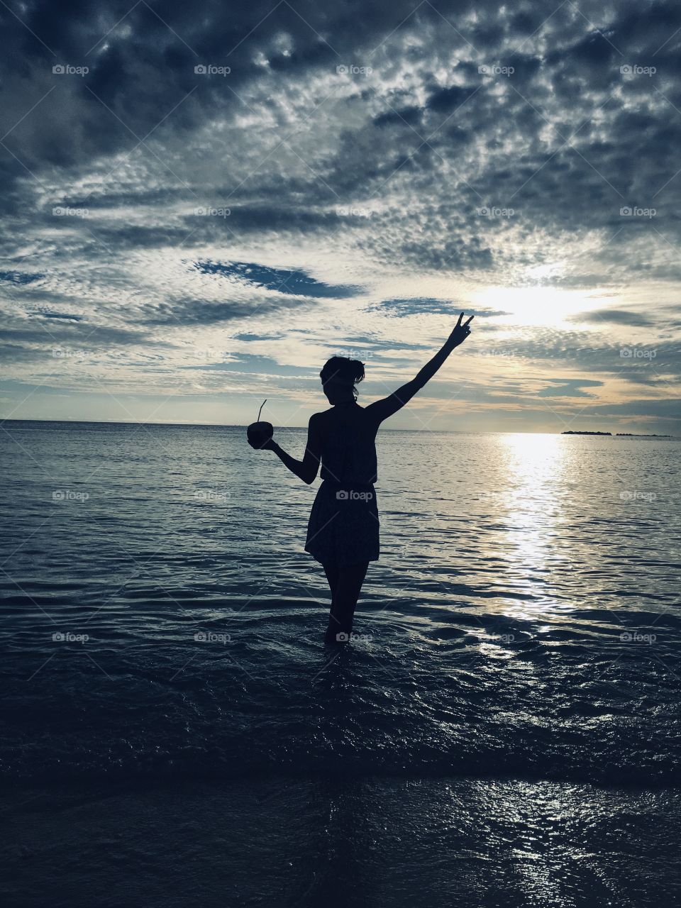 silhouette of a slender girl with a raised hand and a coconut with a straw, against the backdrop of an unusual sunset over the ocean. Maldives, relax