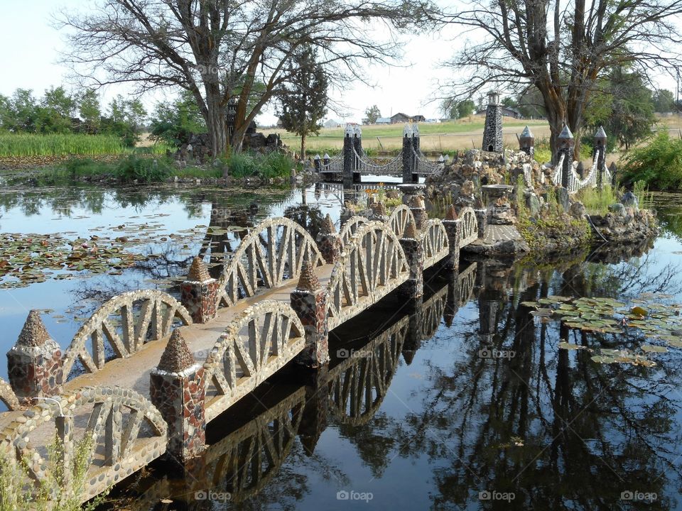 A decorative and detailed rock foot bridge crosses a pond to a center island on a summer day in Central Oregon. 