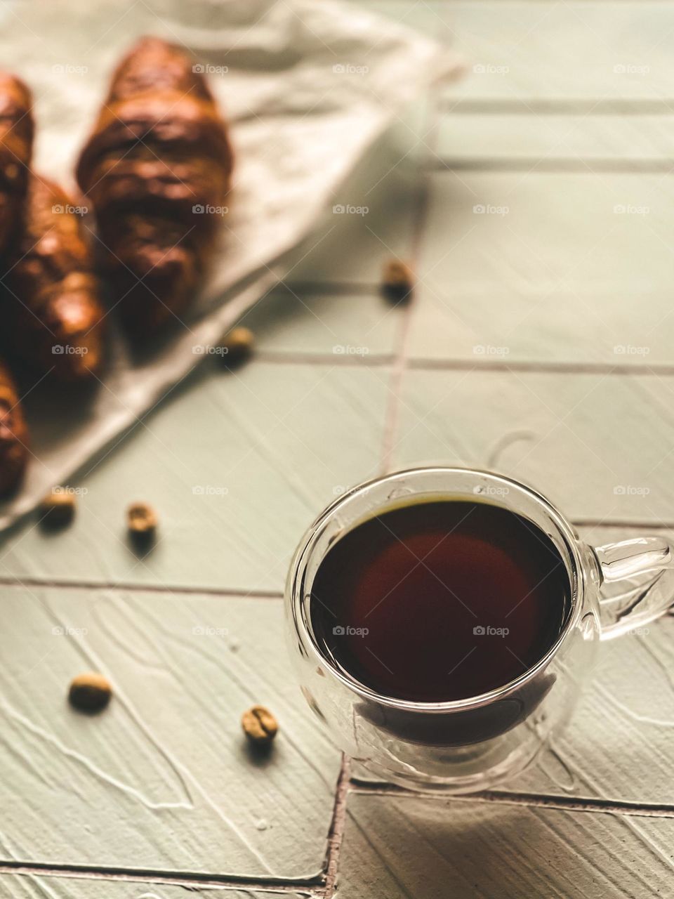 A hot black coffee on a white table top and some croissants on the back. Cold morning