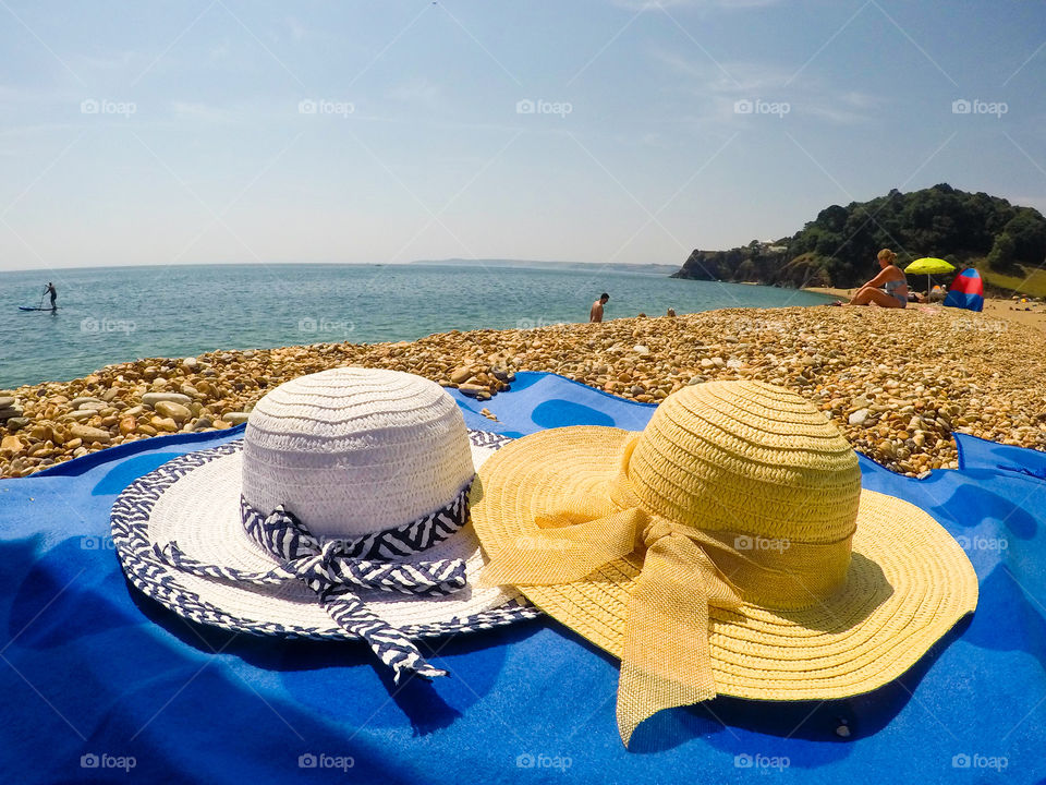 Two hats on top of a blue picnic matt. With pebbles around and a beach at the background.