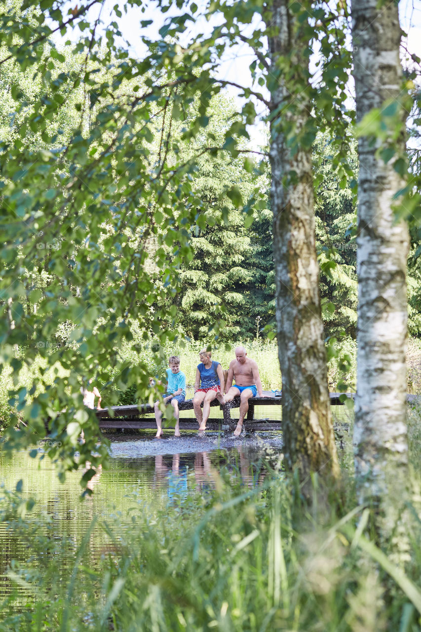 Family spending time together sitting on a bridge over a lake, among the trees, close to nature, during summer vacations. Candid people, real moments, authentic situations