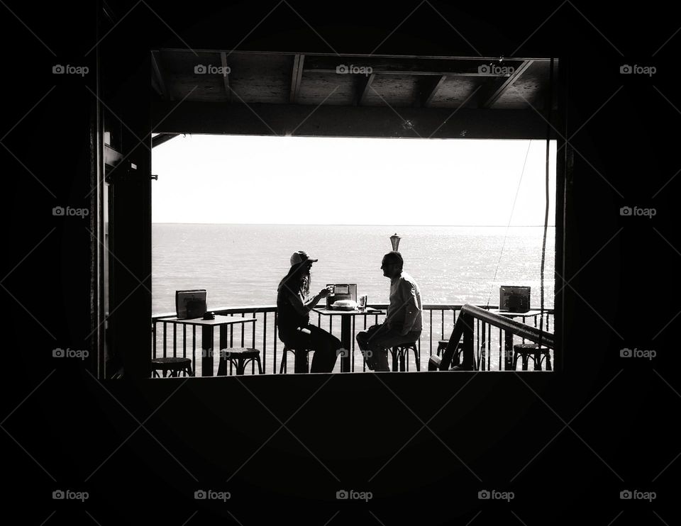 The view through a window of a man & a woman sitting at a table outside by water in Silhouette.