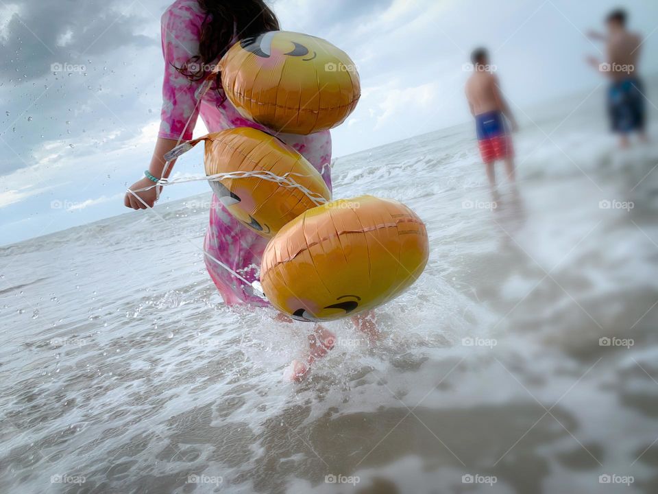 Little Girl Jumping And Running Having Fun With Helium Emoji’s Balloons On The Beach By The Ocean In The Water.