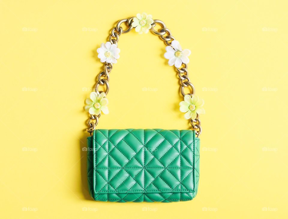 A beautiful stylish green quilted women's handbag with a large gold chain and spring flowers lies in the center on a yellow yon, flat lay close-up. Fashion accessories concept.