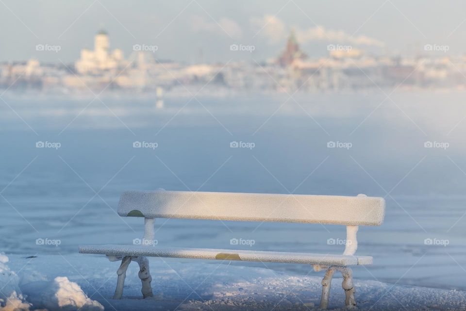 Traditional wooden and frosty resting bench in Helsinki, Finland  on an extremely cold winter morning with freezing Baltic Sea and downtown Helsinki skyline in the background. 
