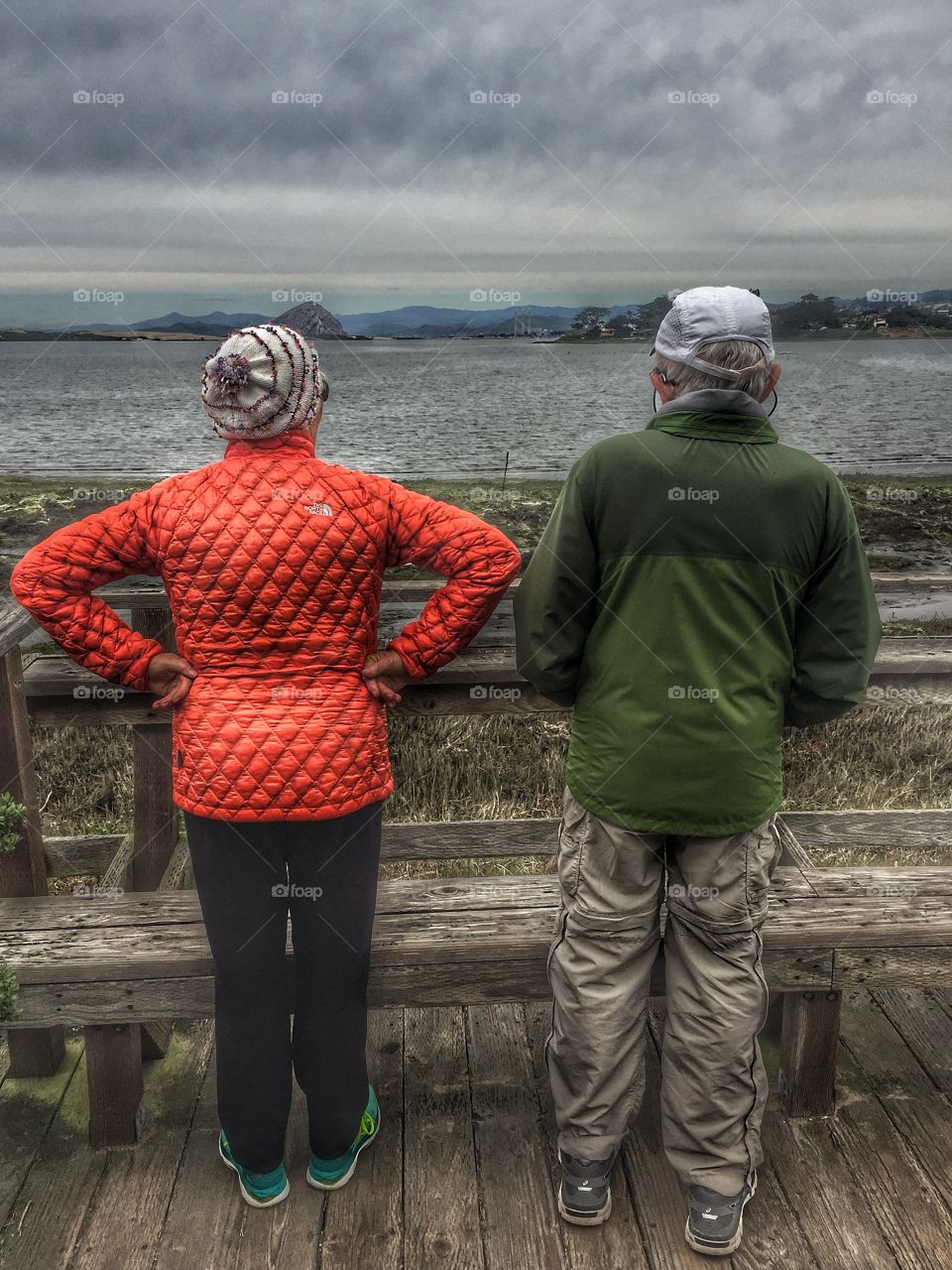 A senior man and woman stand on the landing on a gray and overcast day, admiring the bay with all the birds and ever changing skyline.