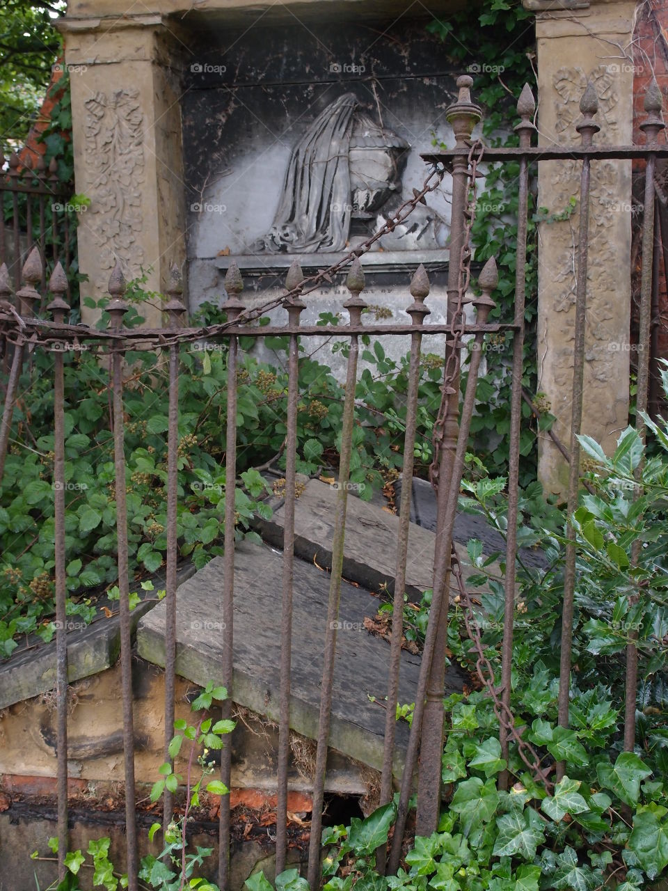 An old European tomb with an iron fence around it, ornate stone carvings, and covered in ivy has withstood the test of time but not without heavy damage. 