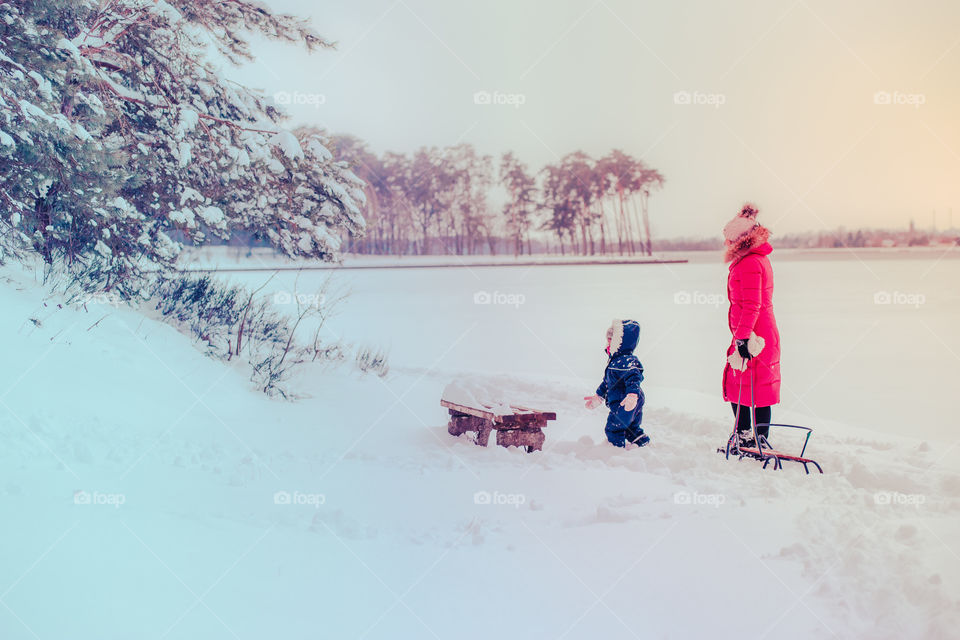 Mother and her little daughter are spending time together walking outdoors in forest in winter while snow falling. Woman is pulling sled, a few years old girl is walking through the deep snow, enjoying wintertime