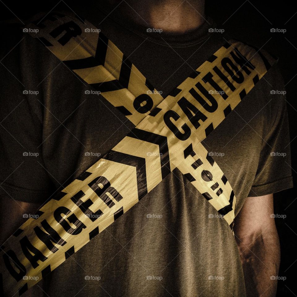 The front torso of man in the dark wearing an olive green T-shirt wrapped in yellow danger/caution tape in the shape of an X. 
Artistic fear symbolism.