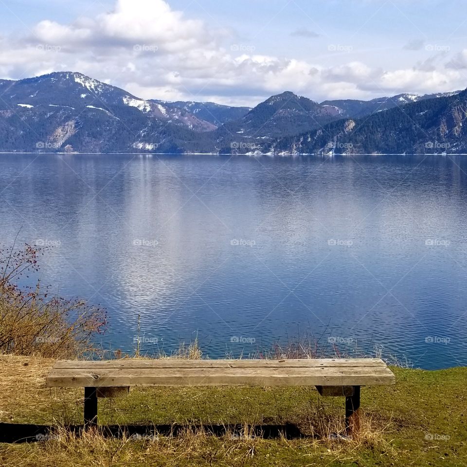 wooden bench with view of  snow covered mountain ridge and reflection on water