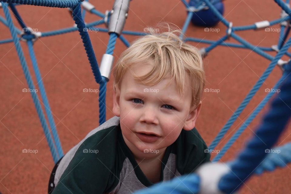 Toddler boy climbing on a bright blue jungle gym at a playground. The boy has short blonde hair.