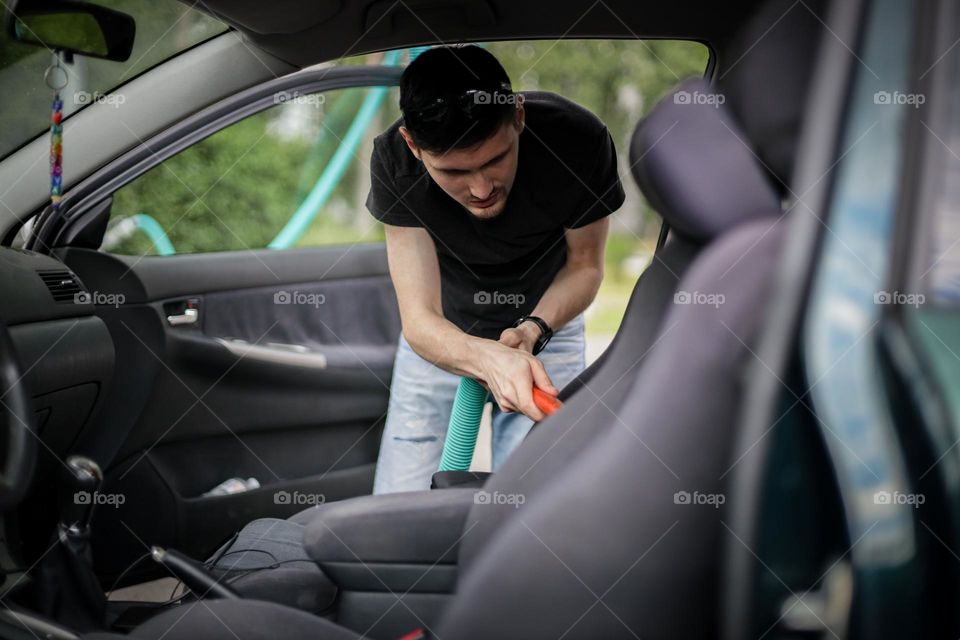 A young handsome caucasian brunette guy in a black t-shirt and light shorts vacuums inside the interior of his car on a summer day at a car wash in Brussels, vtd side close-up. Car wash concept.