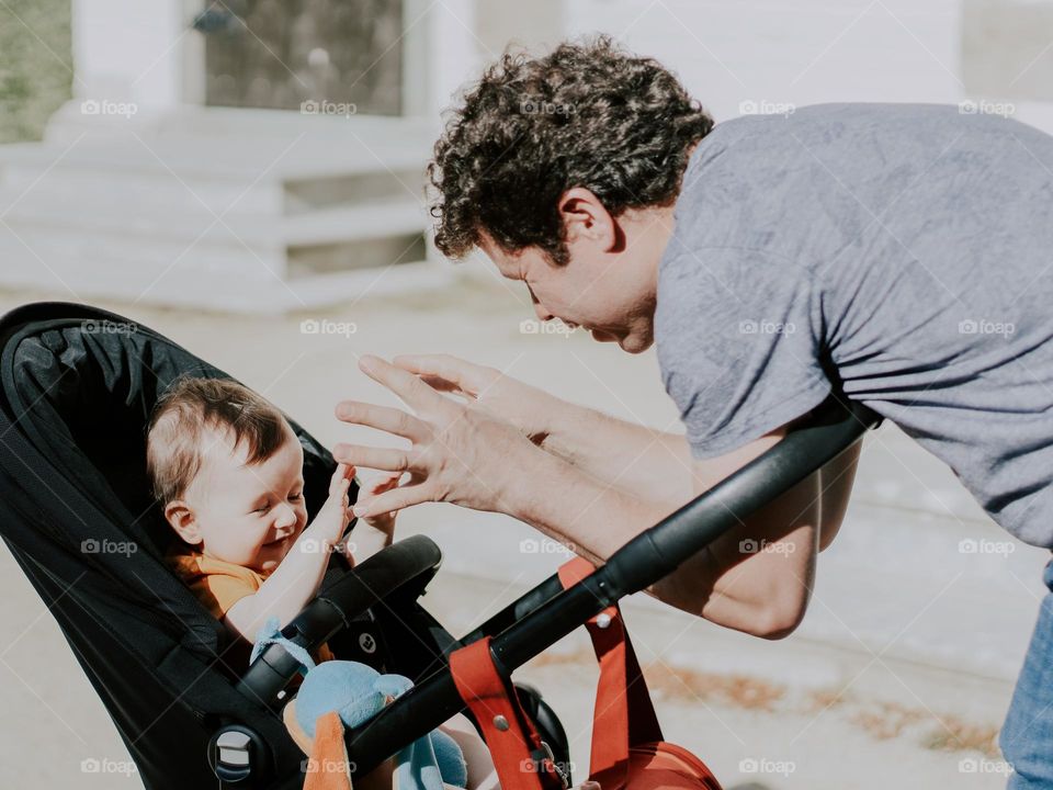 Portrait of young caucasian man with curly brown hair playing with his little daughter slouching in a stroller in a public park on a bright sunny day, close-up side view. Dads concept, family pastime, happy people.