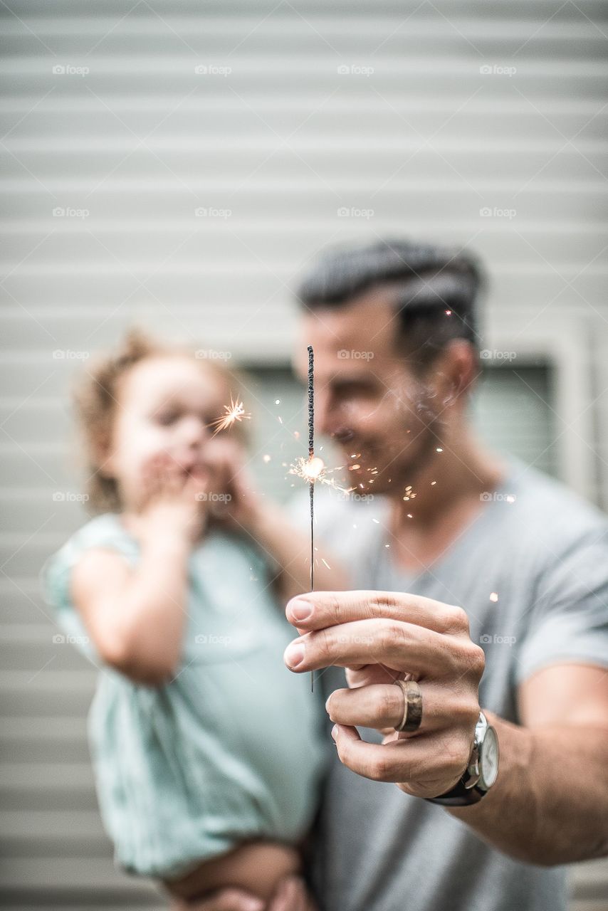 sharply in foreground a father's hand holds up a sparkler as he holds his delighted daughter in other