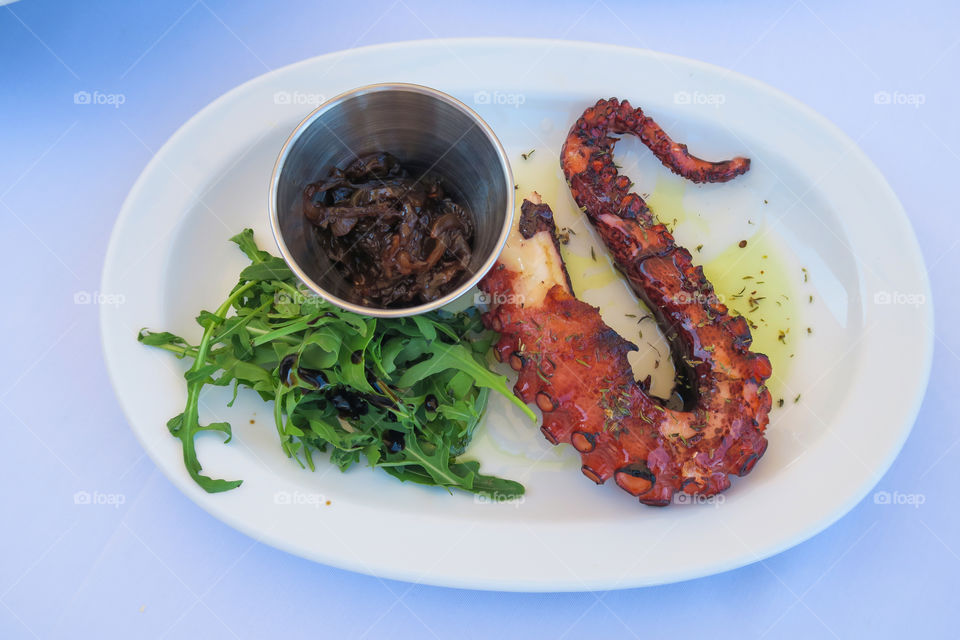 Caramelized octopus with onions and greens, served on a white plate.  Grilled caramelized octopus legs with olive oil on a tavern table by a Greek beach.