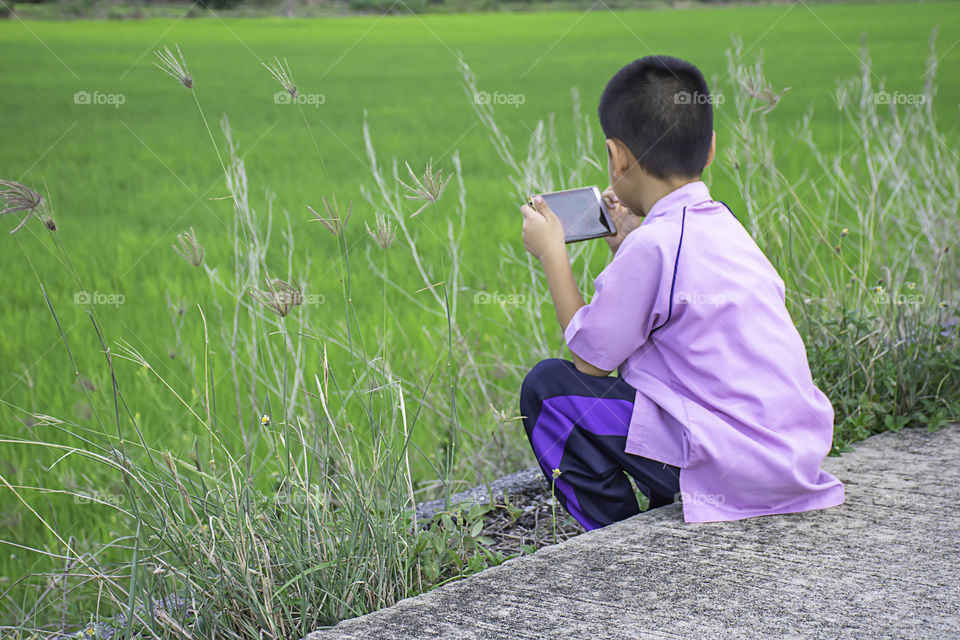 Asian boy holding a phone and sitting on the street  Background the green rice fields.