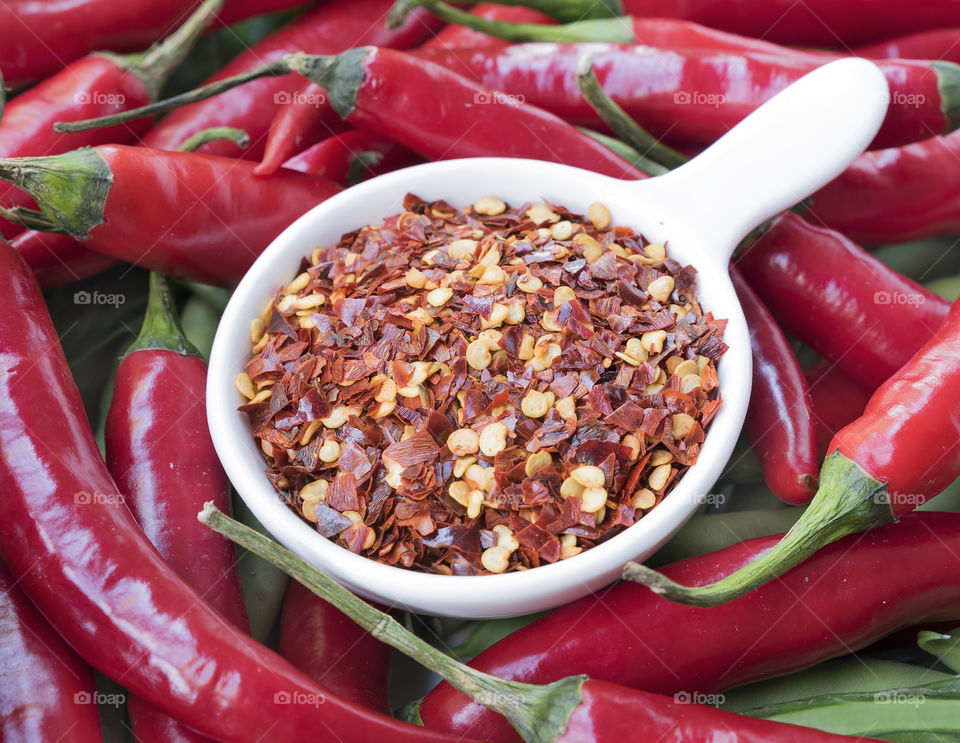 Crushed dried red chilli flakes in a white bowl amongst fresh raw red chillies.