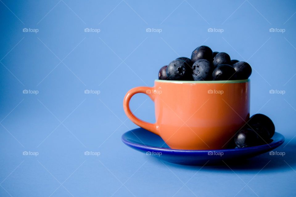 Orange Teacup sitting on Deep Blue saucer filled with Blueberries on Blue background