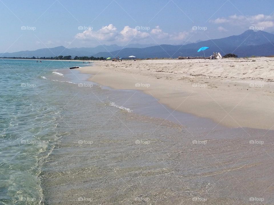 Peaceful Corsican beach in the early morning. The translucent sea and the white sand give the impression of being in paradise. In the background, a bright blue umbrella and the mountains.