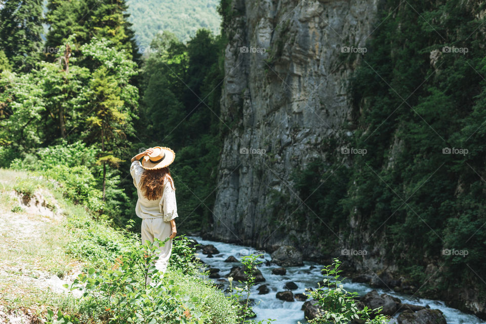Young woman traveler with long blonde hair in straw hat looks at beautiful view of mountain river, people from behind