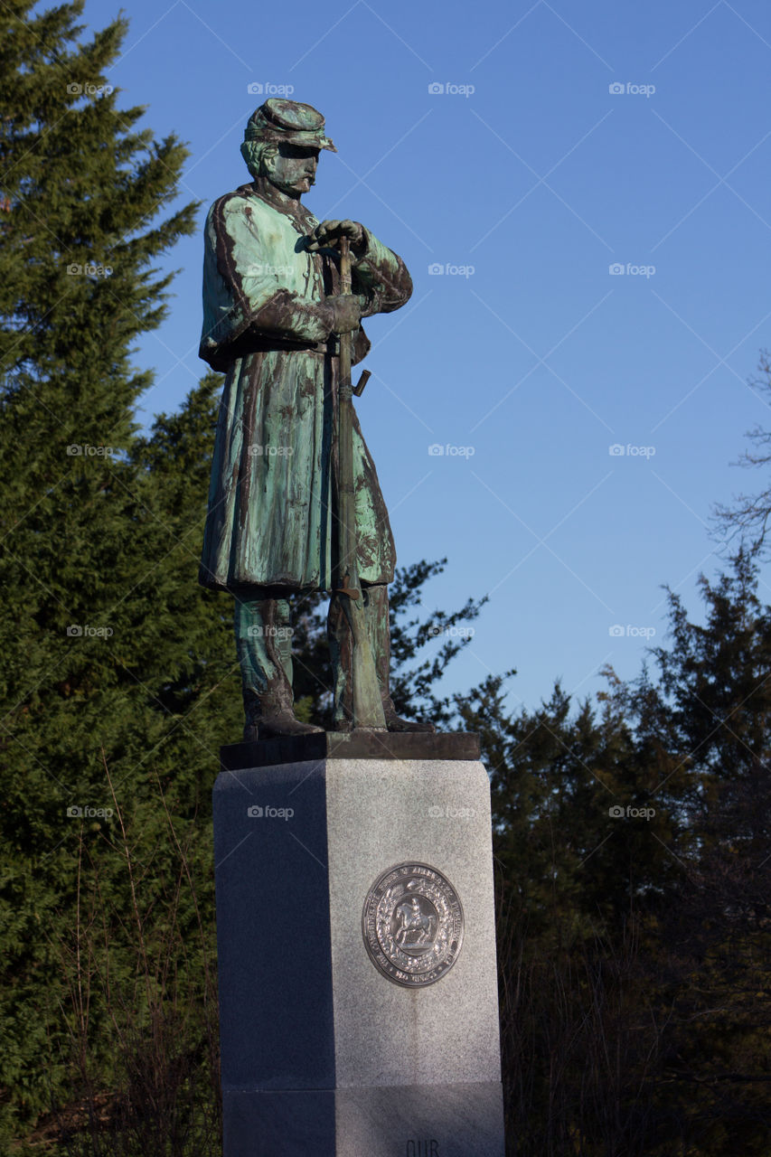 Civil war soldier with great patina on concrete pedestal and blue sky surrounded by pine trees.