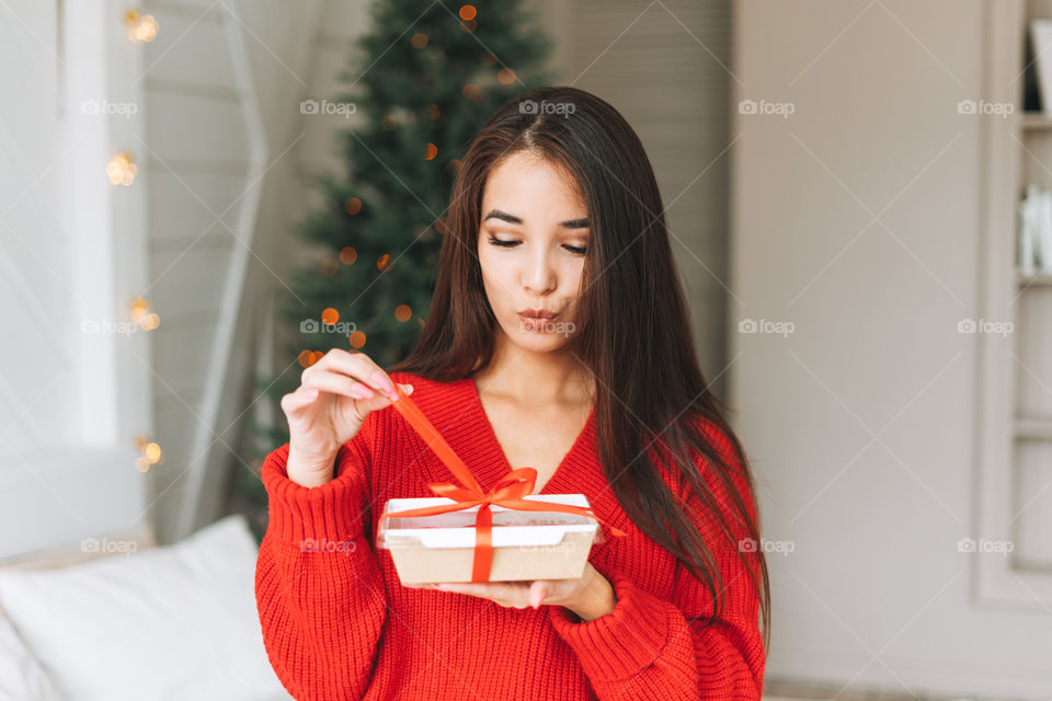Young Asian woman in red sweater with box with candy in hand in bedroom with Christmas tree at home 