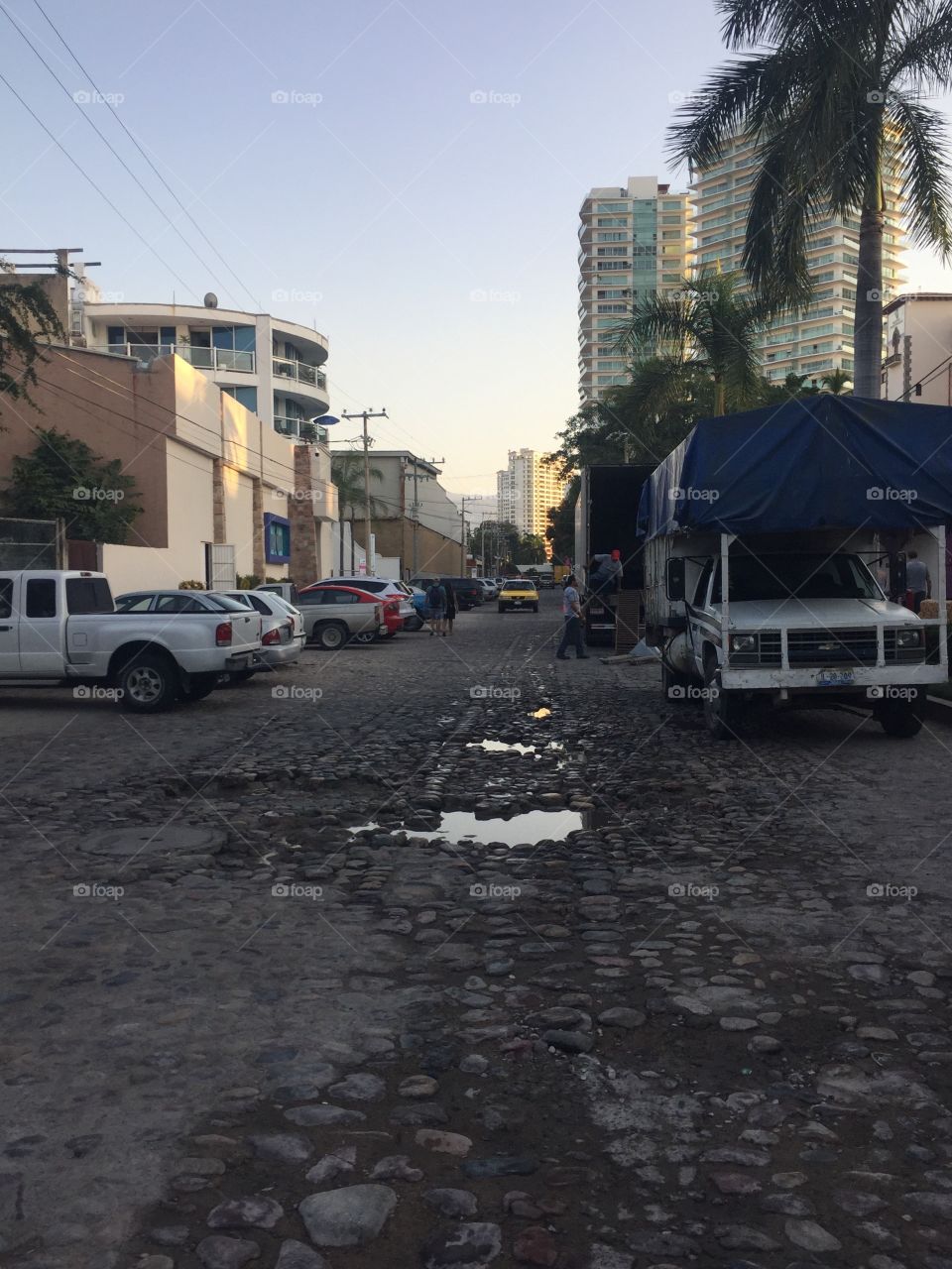 Old cobblestone Road in Puerto Vallarta in the Mexican Riviera with puddles in the middle, cars on the side of the street, and tall, luxury hotels in the distance