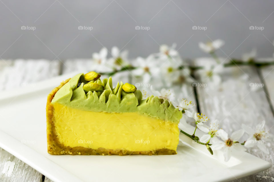Selective focus to piece of mango pistachio cheesecake on white plate at wooden table.