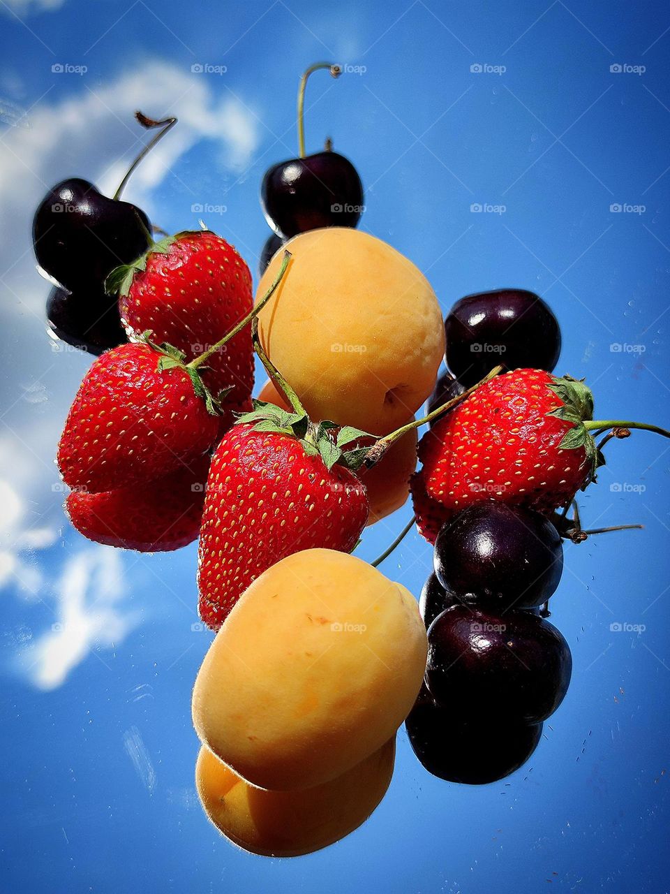 Sunny day. Fruits lie on a mirror surface: orange apricot, red strawberries, dark burgundy cherries against a blue sky with a white cloud. The rays of the sun fall on the fruits and make the fruits juicier