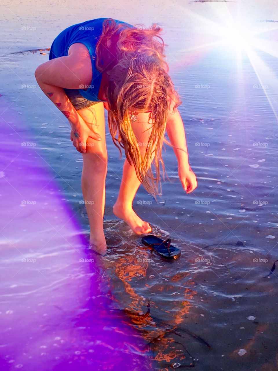 Young ten year old girl with long hair standing in ocean at golden hour at low tide picking up seashell, rainbow light leak