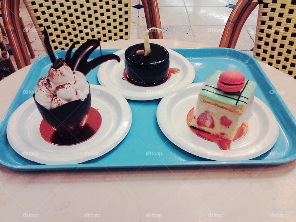 bright blue plastic serving tray with 3 colorful desserts arranged on it. a glossy chocolate cake, a white strawberry cake with a macaron on top and an ice cream sundae with elaborate garnish. in the background is a wicker chair.