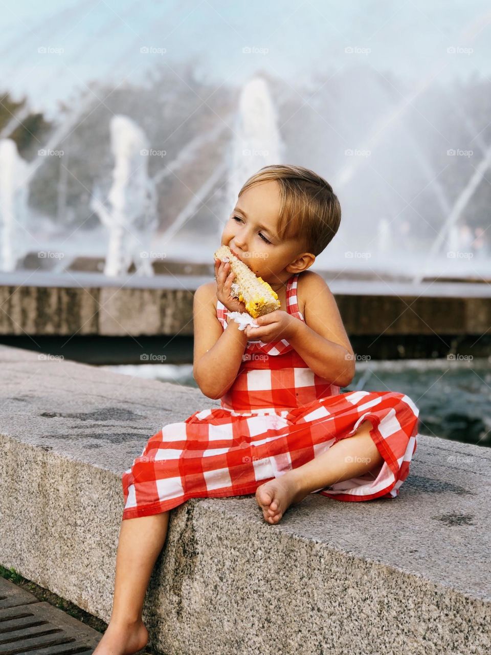 Cute little girl with short blonde hair wearing red dress sitting near fountains and eating corn, portrait of child 