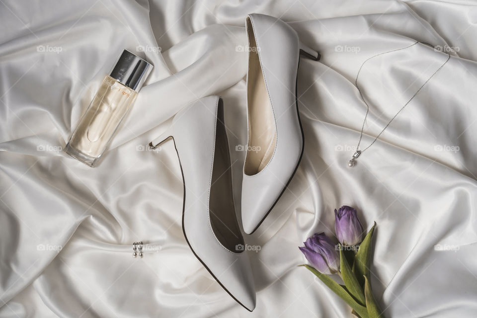 A flatlay of bridal accessories featuring white heels, tulips, jewelry & perfume for a bride on her wedding day 